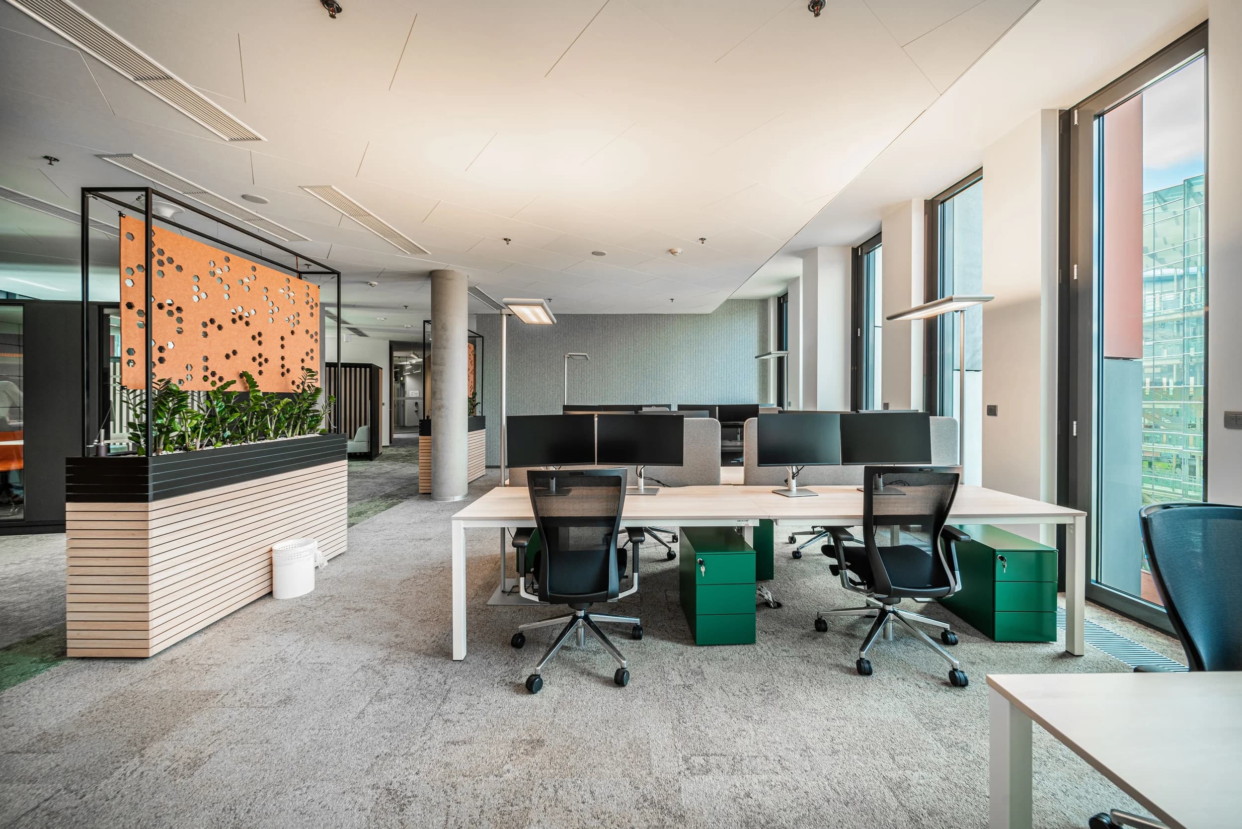 Open space Kooperativy v Praze s pracovními stoly, dvojicí monitorů, zelenými kontejnery a oranžovým paravánem