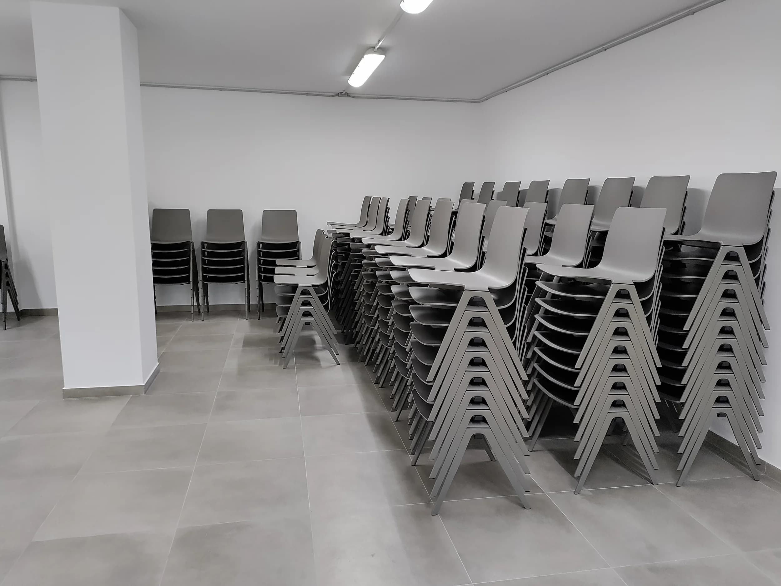 Sloupce šedých skořepinových židlí uskladněné v bílé místnosti s dlažbou, připravené pro multifunkční prostor
